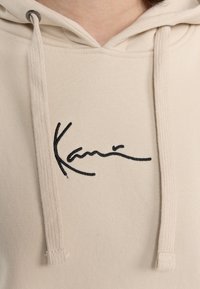 Beige hættetrøje med sort broderet signatur og snore, lavet af blødt, struktureret stof med en afslappet pasform.