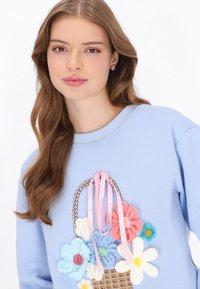 Világoskék pulóver, amelyen egy szőtt kosármintázat látható, színes, kötött virágokkal és rózsaszín szalagdíszítéssel. Puha tapintású.