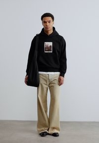 Νέος άνδρας που φοράει μαύρο φούτερ με σχέδιο, μπεζ φαρδιά παντελόνια, μαύρα παπούτσια, και κρατάει μαύρη τσάντα ώμου, στέκεται μπροστά σε λευκό απλό τοίχο.