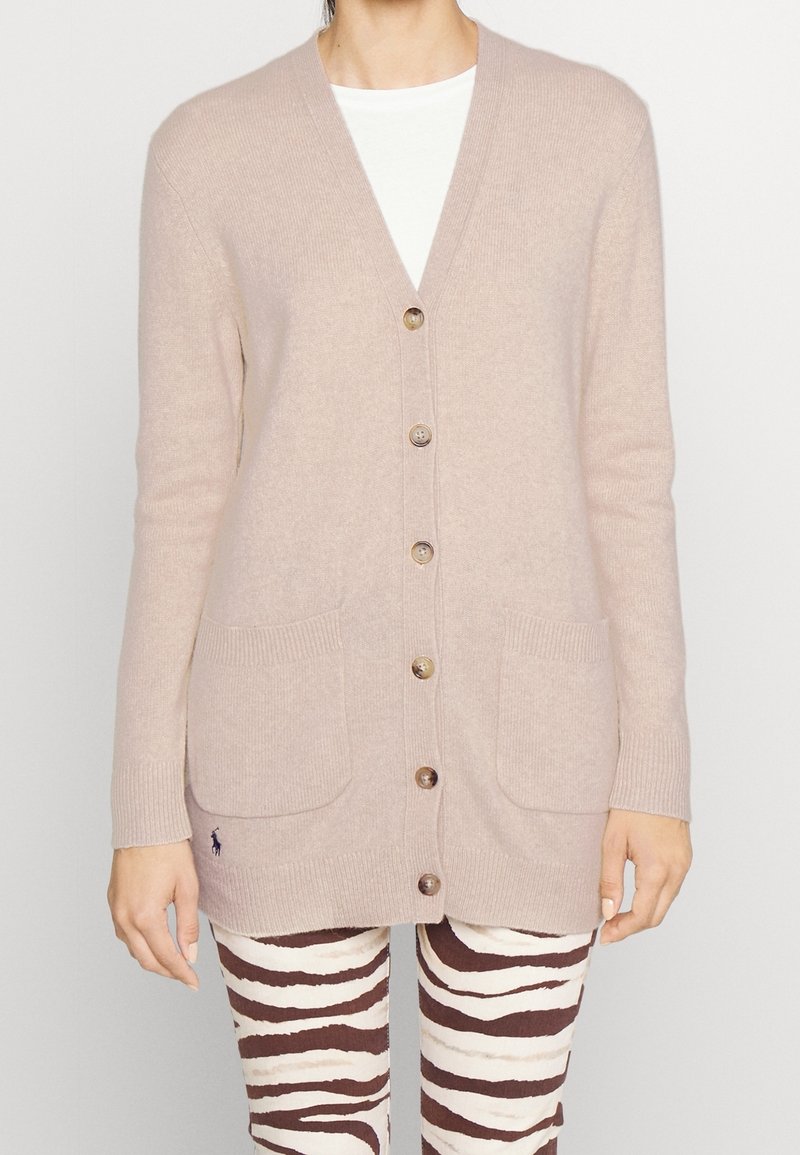 Beige cardigan vyrobený z měkkého materiálu s výstřihem do V, předním zapínáním na knoflíky, dvěma předními kapsami a elastickými manžetami. Výrazné logo značky na dolním lemu.