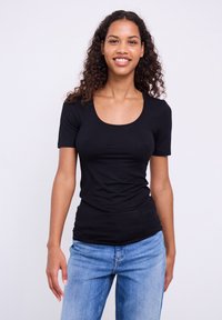 Smilende ung kvinde med krøllet hår iført en tætsiddende sort top med korte ærmer og lyseblå jeans mod en ensfarvet lys baggrund.