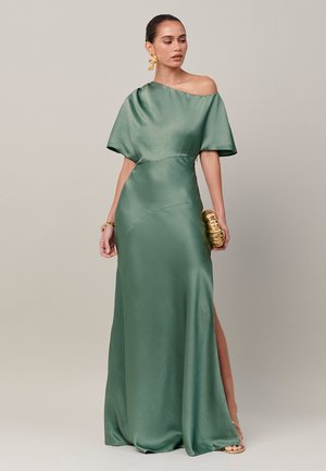 Vrouw draagt een groene satijnen jurk met één schouder en een opgeblazen mouw, houdt een gouden clutch vast en draagt bijpassende gouden oorbellen en armband.