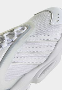 Vitt sportskor med texturerad meshovandel, släta detaljer och sydda sömmar. Skosnörena är fastsatta genom förstärkta öljetter.