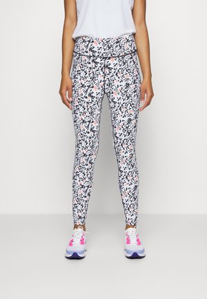 Vrouw draagt een witte legging met hoge taille en een zwart-koraal abstract spettersprint, gecombineerd met witte sneakers met roze accenten.
