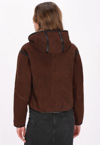 Giacca in suede marrone con cappuccio, caratterizzata da dettagli in metallo nero e cuciture a contrasto. Lunghezza corta, vestibilità rilassata e superficie texturizzata.