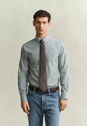 Homme portant une chemise rayée à boutons, une cravate à motifs, une ceinture brune tressée et un jean bleu, debout devant un fond uni clair.