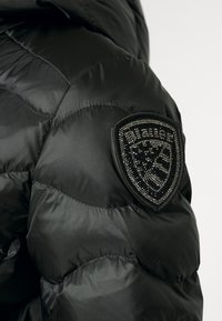 Manche de veste matelassée noire avec un écusson en forme de bouclier orné de perles noires brillantes sur le haut du bras.