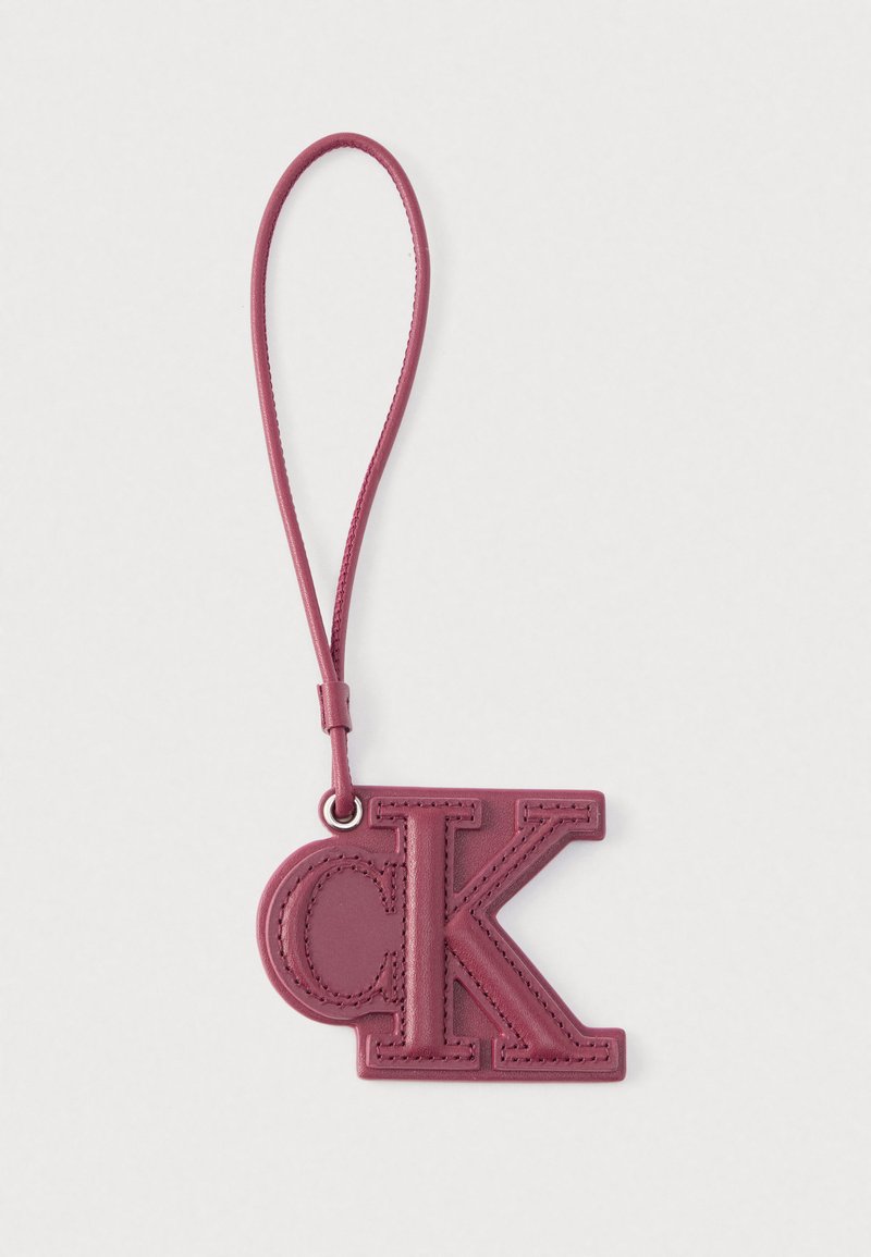 Bracelet de poignet en cuir bordeaux avec un pendentif logo "CK" cousu, attaché par un anneau métallique sur un fond blanc uni.