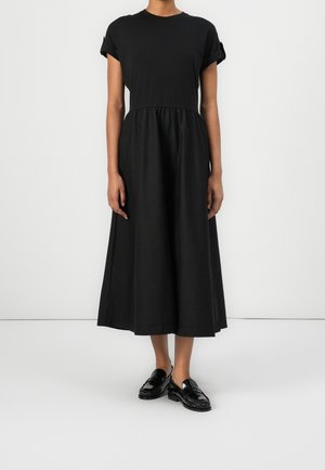 Woman wearing a black, short-sleeve, midi-length dress with a gathered waist and black shiny loafers, standing against a white background.