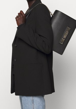 Personne portant un blazer noir et un jean bleu clair, tenant un sac à bandoulière noir Love Moschino avec une chaîne dorée.