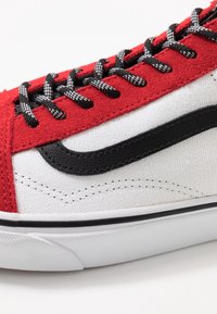 Gros plan d'une basket rouge et blanche avec des lacets noirs et blancs, une bande latérale noire texturée, et une semelle en caoutchouc blanche avec bordure noire.