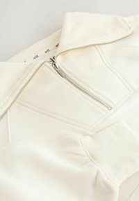 Felpa color avorio con zip e collo alto, tessuto strutturato e dettagli con cuciture diagonali. Presenta una cerniera argentata e polsini a coste.