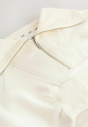 Hanorac alb-crem cu fermoar, guler înalt, material texturat și detalii diagonale ale cusăturilor. Are un fermoar argintiu și manșete tricotate.