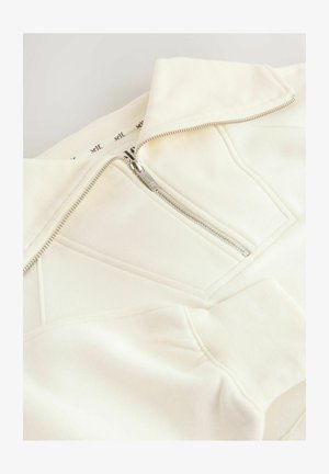 Hanorac alb-crem cu fermoar, guler înalt, material texturat și detalii diagonale ale cusăturilor. Are un fermoar argintiu și manșete tricotate.