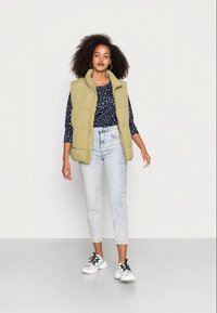 Colete almofadado caqui sobre uma blusa azul-marinho com bolinhas, combinado com jeans de lavagem clara e sapatilhas preto e brancas. A modelo está de pé contra um fundo neutro.