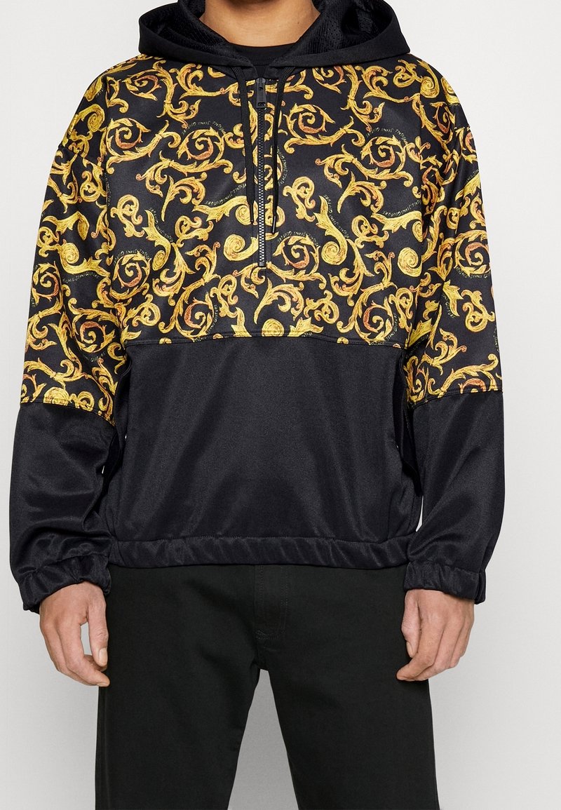 Sweat à capuche avec corps noir et motif baroque doré sur la partie supérieure de la poitrine, les manches et la capuche, avec une demi-fermeture éclair et des poignets élastiques.