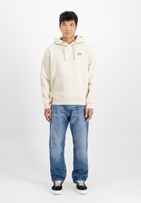 Bežs kapucis ar priekšējo kabatu un aukliņu, ar mazu purpura logotipu uz krūšu daļas. Valkāts ar gaiši ziliem džinsiem un melniem sporta apaviem.