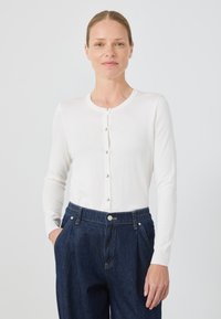 Biały kardigan z długim rękawem, wykonany z teksturowanej dzianiny, z zapięciem na guziki z przodu i srebrnymi guzikami, połączony z ciemnoniebieskimi dżinsami.