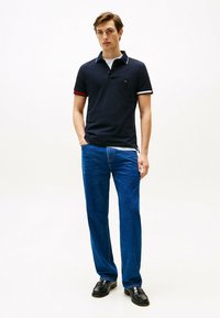 Polo shirt navy con polsini a righe rosse e bianche a contrasto, indossato con jeans blu a gamba dritta e scarpe nere, presenta un piccolo logo sul petto.