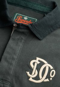 Mørkegrøn polo skjortekrave med hvide broderede, stiliserede initialer og en delvist synlig grøn etiket inde i kraven med teksten "Superdry."