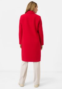 Rode middellange jas met kraag, gemaakt van een gestructureerde stof. Bijpassende crèmekleurige broek met rechte pijpen en beigetinten schoenen.