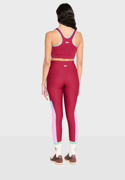 Femme debout de dos, portant un soutien-gorge de sport bordeaux et des leggings avec des panneaux latéraux roses et bleus, des chaussettes blanches et des baskets marron.