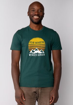Grünes Baumwoll-T-Shirt mit einem Grafikdesign von Bergen, einer Sonne und deutschem Text. Kurze Ärmel und ein Rundhalsausschnitt.