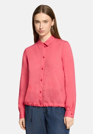 Young woman with straight brown hair wears a bright pink long-sleeve button-up shirt and loose dark blue pants against a plain background.