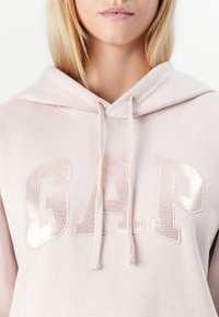 Svijetlo ružičasti dukserica od mekog materijala, s velikim, sjajnim "GAP" logotipom u iridescentnom slogu i vezicom na izrezu.
