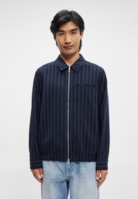 Ναυτικό ζακέτα με φερμουάρ και λευκές κάθετες ρίγες, γιακά, μία τσέπη στο στήθος και ελαστικές μανσέτες; φορεμένη πάνω από ανοιχτό μπλε τζιν παντελόνι.