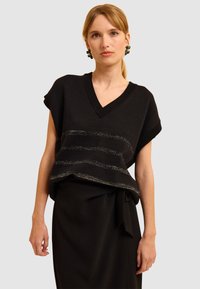 Schwarzer gestrickter Pullover mit V-Ausschnitt, kurzen Ärmeln und horizontalen metallischen Streifendetails. Kombiniert mit einem schwarzen Rock mit Bindegürtel.