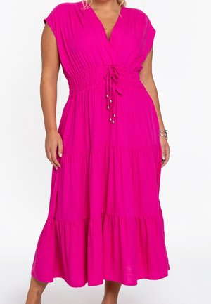 Femme portant une robe midi rose vif avec une taille froncée et une jupe à volants, debout, les mains détendues le long du corps.