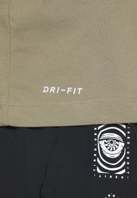 Camisa Dri-FIT verde oliva con detalle del logo en blanco en el dobladillo. Tejido suave y texturizado con un corte casual. Pantalones negros con un estampado gráfico.