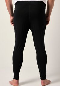 Uomo in piedi a piedi nudi, rivolto di spalle, che indossa leggins neri aderenti e una maglietta bianca, su uno sfondo neutro e semplice.