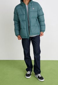 En teal pufferjacka med quiltat mönster, framsidan med dragkedja och huva, bärs över en ljusgrå skjorta och kombineras med mörka jeans och svarta sneakers.