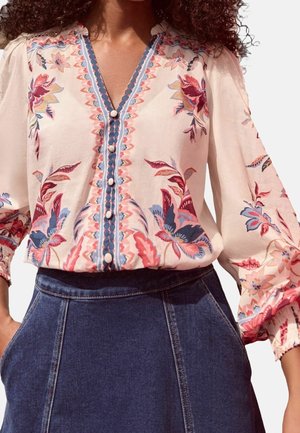 Femme portant un chemisier à motifs floraux avec des boutons blancs et une jupe en jean bleu foncé taille haute, les mains dans les poches.