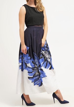 Woman wearing a sleeveless black top and a high-waisted maxi skirt with large blue and white floral patterns, paired with navy high heels.