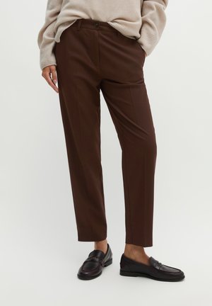 Person wearing dark brown straight pants, beige sweater, and dark brown loafers standing with one hand in pocket.