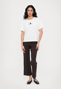 Weißes Baumwoll-T-Shirt mit kurzen Ärmeln, das mit einem schwarzen Grafikdesign versehen ist. Kombiniert mit braunen Schlaghosen und schwarzen flachen Schuhen.