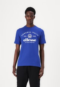 T-shirt de algodão azul com decote redondo e mangas curtas. Apresenta texto branco com o nome da marca e impressões de localização na frente.