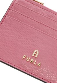 Růžová texturovaná kožená peněženka na karty se čtyřmi sloty na karty, zlatým logem a zipovým uzávěrem nahoře. Viditelné akcentované švy.