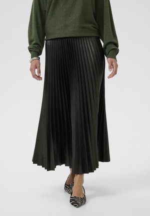 Schwarzer plissierter Rock aus Kunstleder, mit einem langen Design und vertikalen Falten; kombiniert mit einem grünen Pullover und gemusterten Absatzschuhen.