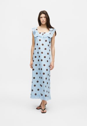 Jeune femme debout, portant une robe maxi bleu clair sans manches à gros pois marron, décolleté carré à volants et sandales noires.