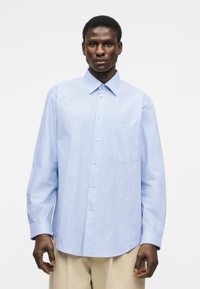 Άνδρας με γαλάζιο πουκάμισο με κάθετες ρίγες και τσέπη, συνδυασμένο με μπεζ παντελόνι, στέκεται μπροστά σε λευκό φόντο.