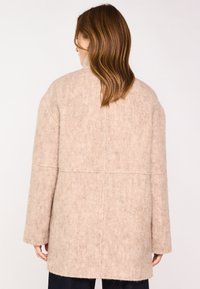 Cappotto in lana beige con una vestibilità rilassata, caratterizzato da un colletto alto, spalle calate e una texture morbida. Le cuciture distintive creano un design minimalista.