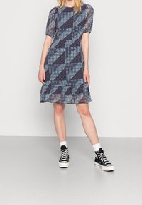 Blau gemustertes Kleid mit kurzen Puffärmeln, gerüschtem Saum und geometrischen Mustern. Kombiniert mit schwarzen High-Top-Sneakers und weißen Socken.