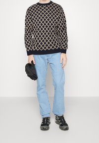 Maglione blu navy con motivo geometrico beige a contrasto, jeans azzurri e sneakers scure. Tiene una piccola borsa nera.