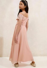 Lichtroze jurk met off-shoulder mouwen versierd met kralen, vloeiend materiaal en een strik om de taille, gecombineerd met glinsterende instap schoenen.