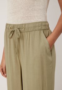 Oliwkowo zielone, lekkie spodnie o długości 7/8 z regulowanym sznurkiem w pasie, bocznymi kieszeniami i gładką fakturą. Elastyczny pas z ozdobnymi troczkami.