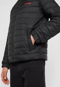 Veste noire matelassée avec logo "HUGO" rouge sur la poitrine, main dans la poche latérale à bouton-pression, portée avec un pantalon noir.